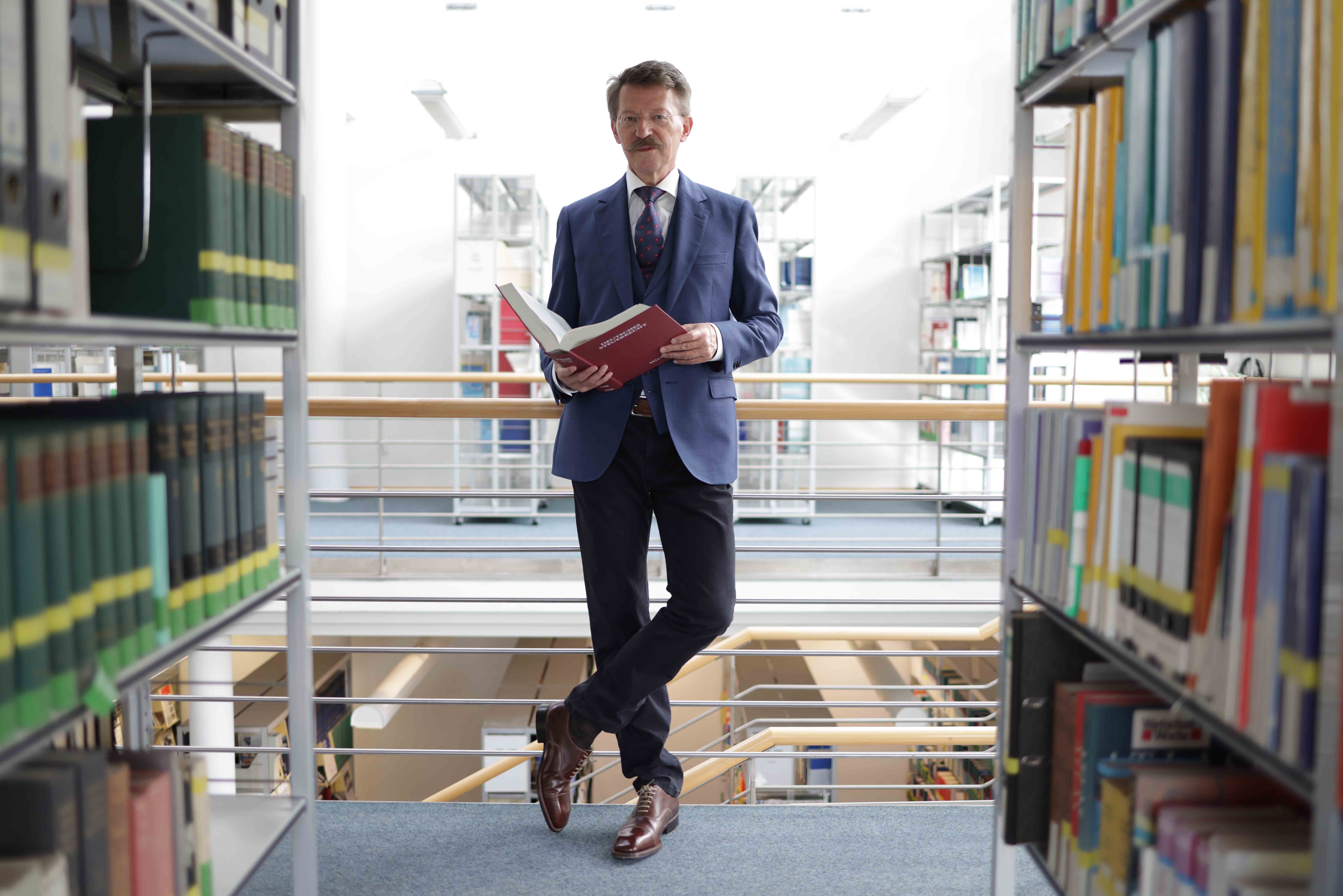 Prof. Köster steht zwischen Bücherregalen mit einem aufgeklappten Buch