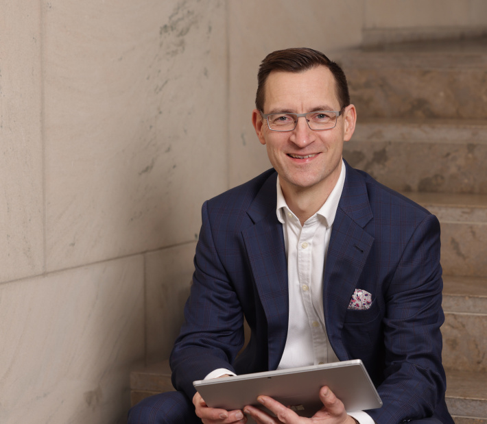 Portrait von Prof. Dr. Mike Schneider schaut lächelnd in die Kamera, sitzt auf einer Treppe und hält ein Tablet in der Hand