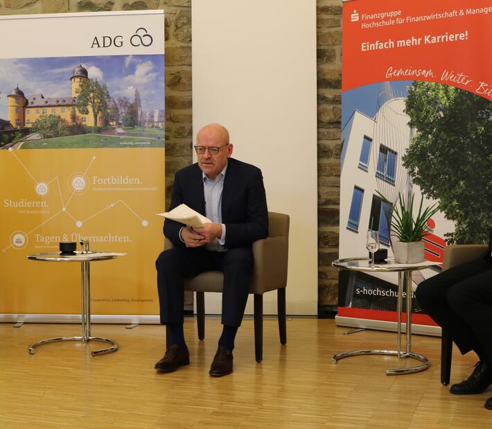 Eine Frau mit Notizblock und zwei Männer im Anzug sitzen beisammen und schauen ins Publikum