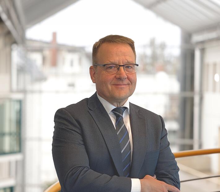 Maik Grabau, in Anzug und Krawatte, steht mit verschränkten Armen vor einer modernen Architektur.