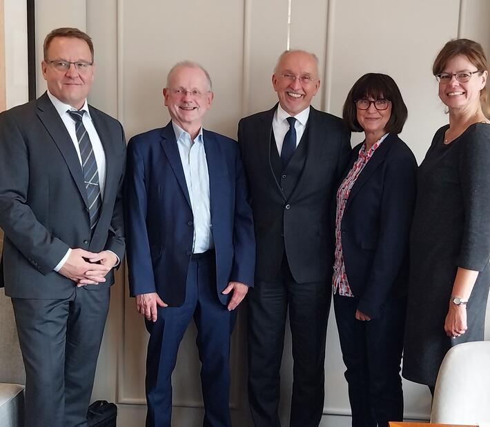 5 Personen in Business Kleidung stehen vor einer Wand und lächeln