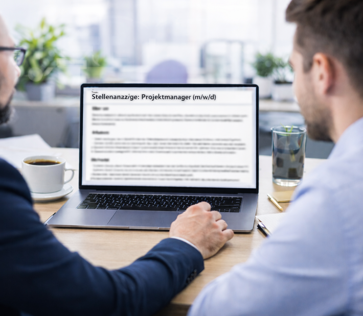 Zwei Männer schauen auf einen Monitor, der eine Stellenausschreibung zeigt.