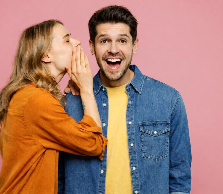 Junge Frau pflüstert jungem Mann etwas ins Ohr, sodass der Mann erfreut strahlt.