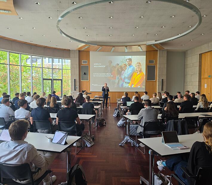 Blick in die Aula der Hochschule mit sitzenden jungen Menschen und dem Rektor