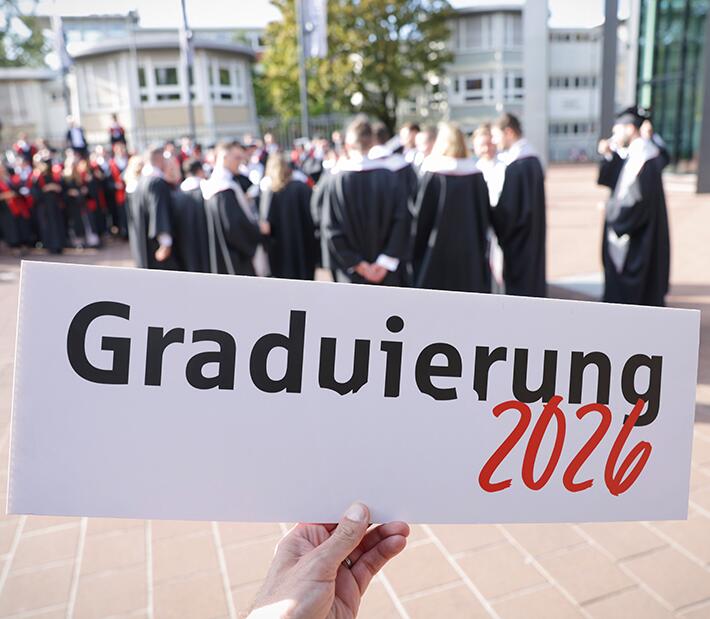 Im Vordergrund ein Schild, auf dem steht: Graduierung 2026. Dahinter stehen Absolventen in Talaren.