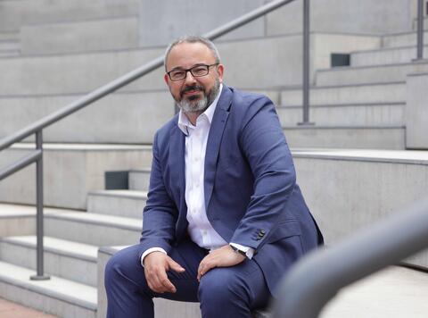 Prof. Dr. Denis Basi sitzt auf einer Treppe, trägt einen blauen Anzug und schaut freundlich in die Kamera.