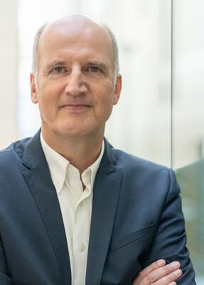 Prof. Dr. Tobias Engelsleben steht an einer Glaswand mit verschränkten Armen und blickt in die Kamera.