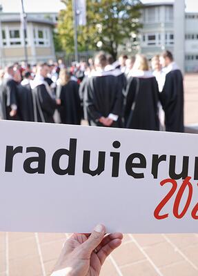 Im Vordergrund ein Schild, auf dem steht: Graduierung 2026. Dahinter stehen Absolventen in Talaren.