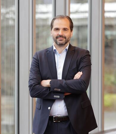 Prof. Dr. Martin Scheffel steht mit verschränkten Armen vor großen Fenstern in einem modernen Gebäude.