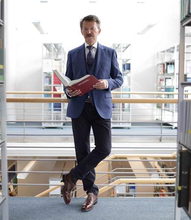 Prof. Dr. Thomas Köster steht in einer Bibliothek und hält ein Buch in der Hand.