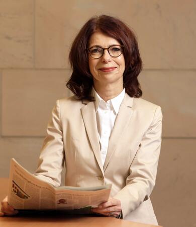 Anne Böhm-Dries sitzt an einem Tisch und hält eine Zeitung in der Hand. Sie trägt einen hellen Blazer und ein weißes Hemd.