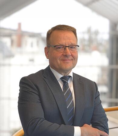 Maik Grabau, in Anzug und Krawatte, steht mit verschränkten Armen vor einer modernen Architektur.