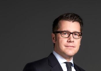 Mann mit Brille und Anzug, lächelnd und im Portraitstil vor grauem Hintergrund.