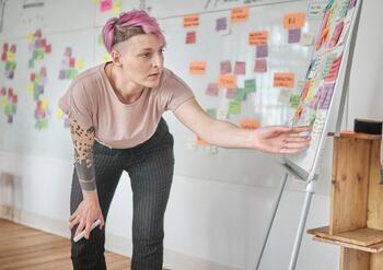 Frau mit pinken Haaren zeigt auf ein Flipchart in einem Meetingraum mit Post-its.