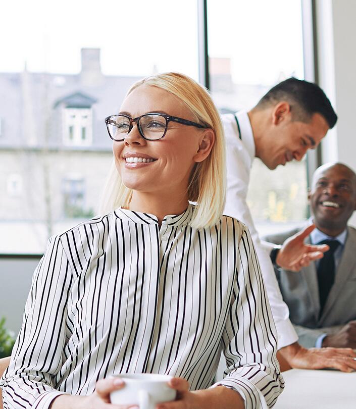 Frau mit Brille lächelt und hält eine Tasse in einem modernen Büro, im Hintergrund sind zwei Männer beim Gespräch.
