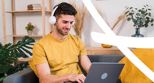 junger Mann im gelben T-Shirt sitzt auf der Couch mit Kopfhörern und lernt am Laptop