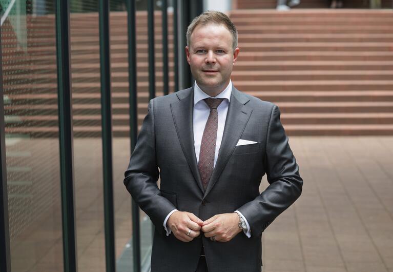 Prof. Dr. Patrick Kuchelmeister in Anzug steht vor Gebäudefassade mit Glas und Treppenanlage.