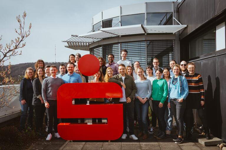 Gruppe von 24 Personen vor einem großen roten Logo in einem Außenbereich.