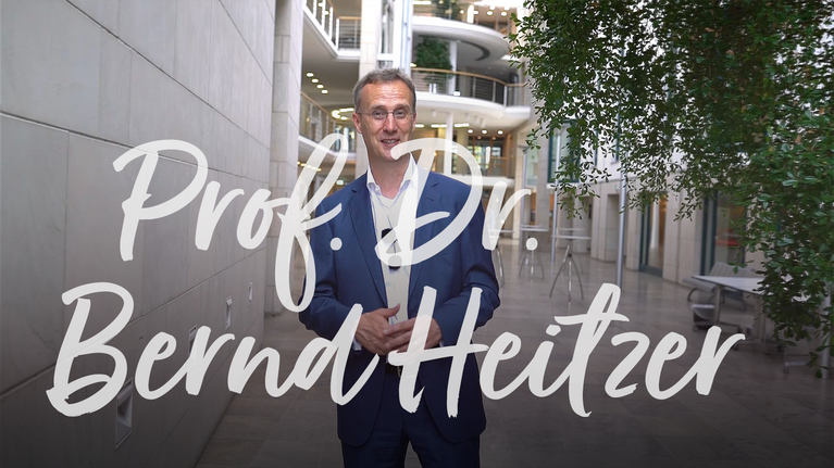 Professor Bernd Heitzer steht im Flur eines modernen Gebäudes und hält die Hände vor sich.