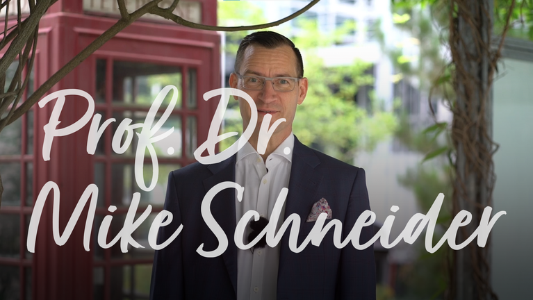 Prof. Dr. Mike Schneider steht lächelnd vor einer grünen Umgebung, bekleidet mit einem Anzug und einer weißen Bluse.