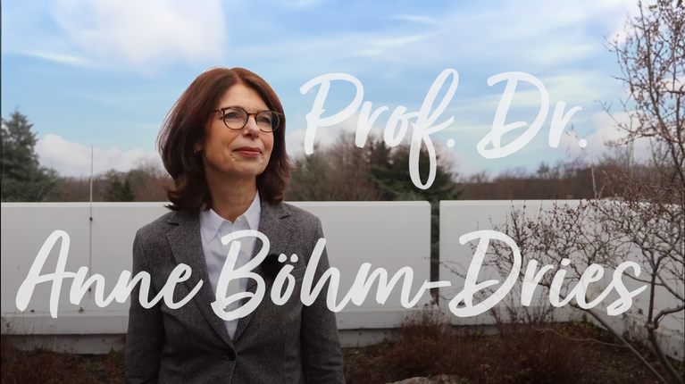 Prof. Dr. Anne Böhm-Dries steht im Freien vor einer weißen Wand, umgeben von Bäumen und Sträuchern.