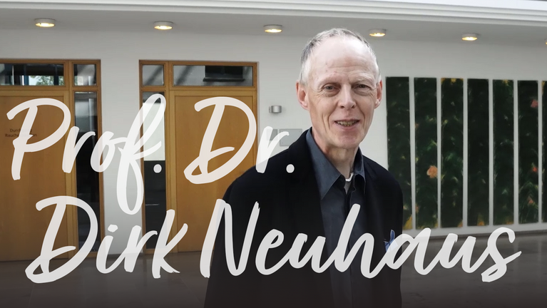 Prof. Dr. Dirk Neuhaus steht an einer Wand mit Pflanzenmotiven und spricht.