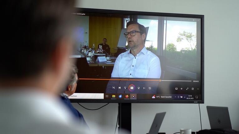 Mann spricht in Video zu Teilnehmern eines Meetings, Monitor zeigt Interviewausschnitt.