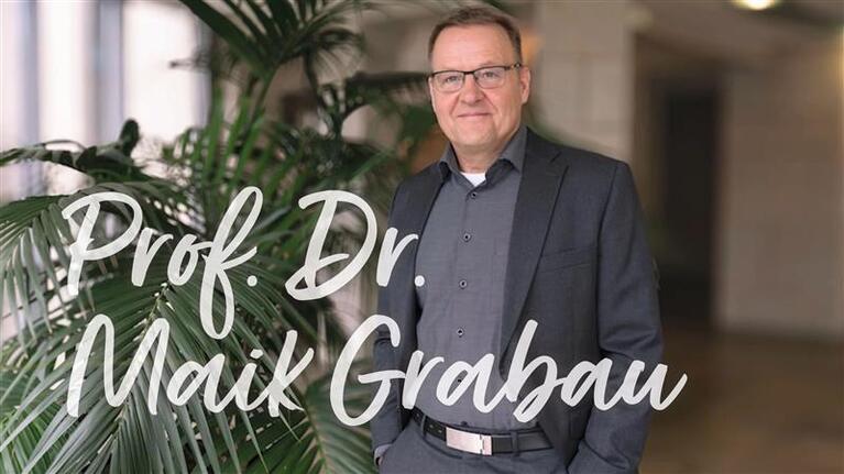 Prof. Dr. Maik Grabau steht vor einer Pflanze in einem Gebäude.